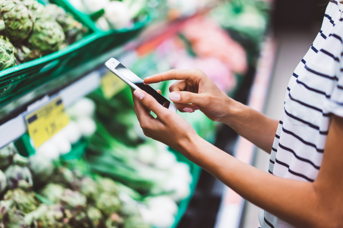 Young woman shopping purchase healthy food in supermarket blur background. Close up view girl blogger buy products using smartphone in store. Hipster at grocery using smartphone. Person comparing price